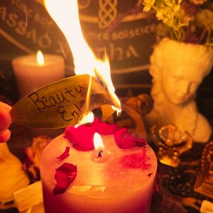 May include: A lit candle with red rose petals and a burning leaf with the words "Beauty En" written on it. A small statue of a woman's head and a small candle are in the background. The theme is spiritual.