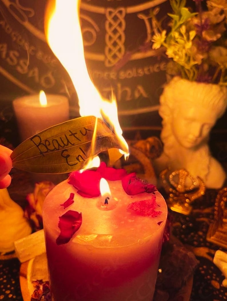 May include: A lit candle with red rose petals and a burning leaf with the words "Beauty En" written on it. A small statue of a woman's head and a small candle are in the background. The theme is spiritual.
