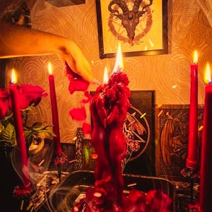 May include: A ritual scene featuring a large red candle shaped like two figures embracing, surrounded by burning red candles and rose petals. A framed image of a goat's head is visible in the background, along with other occult symbols.