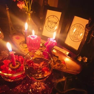 May include: A dimly lit scene featuring lit candles, a glass of wine with flower petals, and framed illustrations. A small Eiffel Tower replica and a vase of flowers are also visible. The overall aesthetic suggests a mystical or spiritual theme.