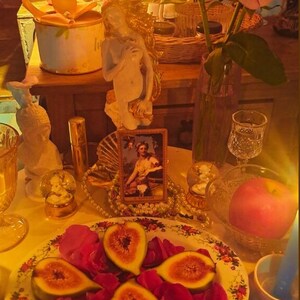 May include: A still life arrangement featuring a plate of figs and rose petals, a vase of roses, candles, and decorative items. The scene is illuminated by warm light, creating a cozy and inviting atmosphere. A small framed picture and a pearl necklace are also present.