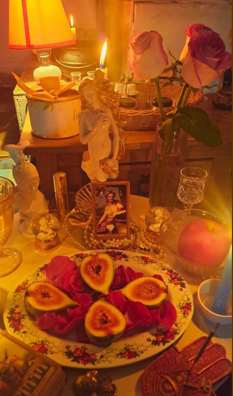 May include: A still life arrangement featuring a plate of figs and rose petals, a vase of roses, candles, and decorative items. The scene is illuminated by warm light, creating a cozy and inviting atmosphere. A small framed picture and a pearl necklace are also present.