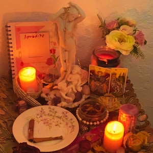 May include: A spiritual altar featuring a white Aphrodite statue, lit candles, tarot cards, and a notebook with the word "Aphrodite." A plate with cinnamon sticks and dried flowers is also present. The scene is illuminated by warm, soft lighting.