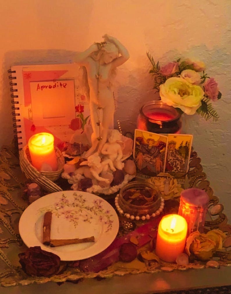 May include: A spiritual altar featuring a white Aphrodite statue, lit candles, tarot cards, and a notebook with the word "Aphrodite." A plate with cinnamon sticks and dried flowers is also present. The scene is illuminated by warm, soft lighting.
