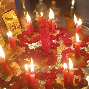 May include: A mystical scene featuring lit red candles arranged with red rose petals on a reflective surface. Several glass bottles and a book with the text "Caggia practica" are visible, creating a spiritual ambiance.