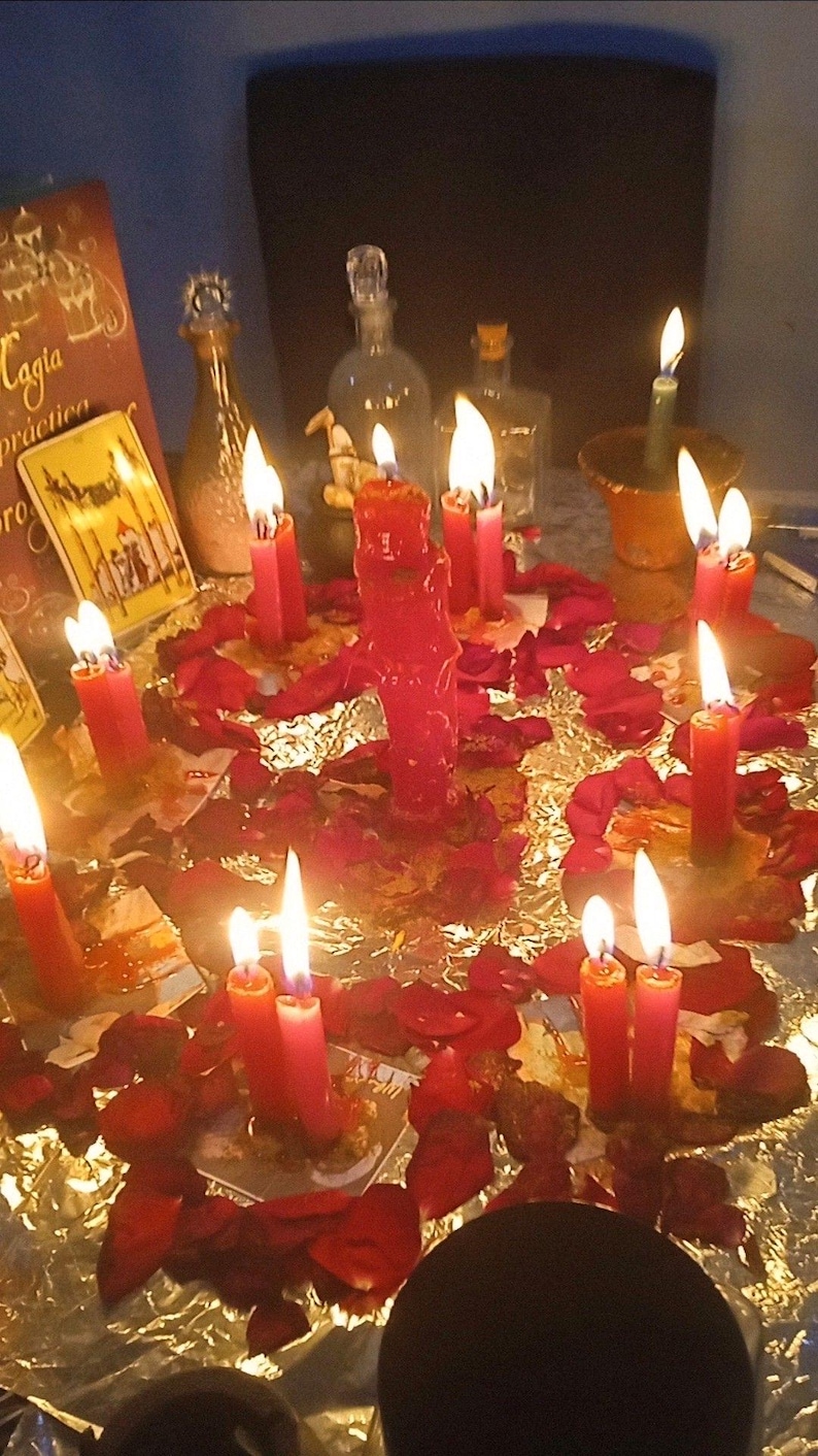 May include: A mystical scene featuring lit red candles arranged with red rose petals on a reflective surface. Several glass bottles and a book with the text "Caggia practica" are visible, creating a spiritual ambiance.