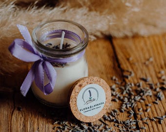 May include: A small glass jar candle with a purple ribbon and a cork lid. The lid has a label that says "Flora & Lavanda L'essenza dalla natura". Dried lavender flowers are scattered around the candle.