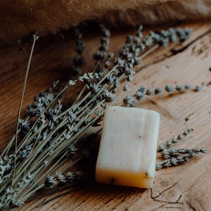 Può includere: Una saponetta rettangolare fatta a mano con una tonalità giallo chiaro, appoggiata su una superficie di legno. Rami di lavanda secca ed erba della pampa beige e soffice sono disposti attorno al sapone, creando un'estetica naturale simile a quella di una spa.