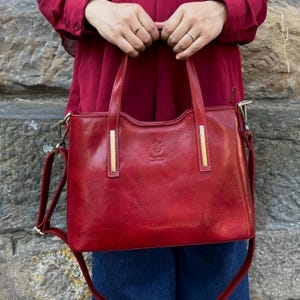 Op de afbeelding: Een rode leren handtas met goudkleurige accenten. De tas heeft een handvat aan de bovenkant en een afneembare schouderriem. De tas wordt vastgehouden door een persoon die een rood shirt en een blauwe spijkerbroek draagt. De tas heeft een klein reliëf op de voorkant.