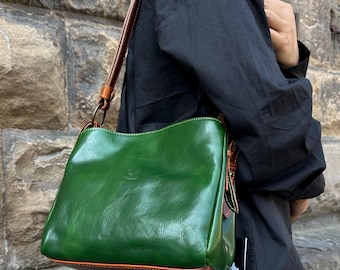 May include: A green leather shoulder bag with brown trim and a matching strap. The bag has a glossy finish and a simple, classic design. The bag is carried by a person wearing a black shirt.