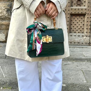 Peut inclure: Un sac à main vert foncé avec une texture peau de crocodile, des poignées en bambou et un fermoir doré. Un foulard à motifs verts, blancs et roses est noué à la poignée. Le sac est tenu par une personne portant un trench-coat beige et un pantalon blanc.