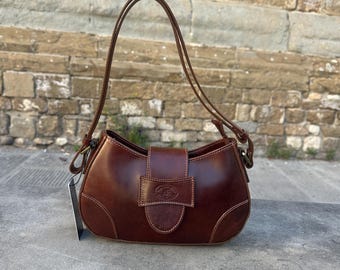 May include: A brown leather handbag with a curved shape and a top handle. The bag features a flap closure with a buckle and a stitched design. The bag is sitting on a concrete surface.