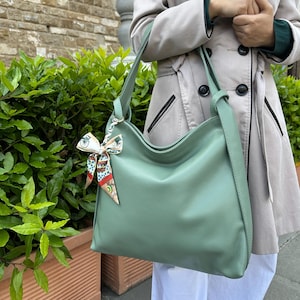 May include: A pale green shoulder bag with a long strap. The bag is made of a smooth material and has a decorative scarf tied to the strap. The bag is being held by a person wearing a light-coloured coat.