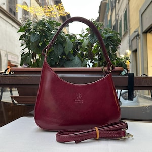 May include: A burgundy leather handbag with a curved shape and a shoulder strap. The bag has a top handle and a detachable crossbody strap. The bag has a small logo on the front and gold-colored hardware. The bag is sitting on a white surface.