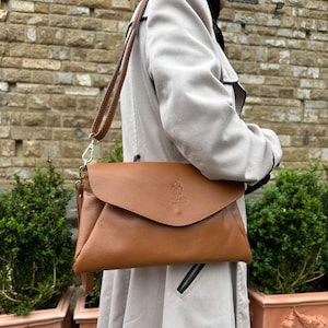 May include: A brown leather crossbody bag with a flap closure and a long adjustable strap. The bag has a textured surface and a small embossed design on the front. The bag is being worn over a light-colored coat.