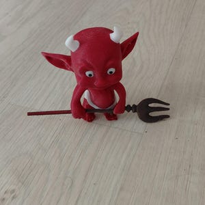 Peut inclure: Une petite figurine rouge de diable avec des cornes et des yeux blancs, tenant un trident marron foncé. Le personnage possède des oreilles pointues et une finition mate lisse, se tenant debout dans une position orientée vers l'avant.