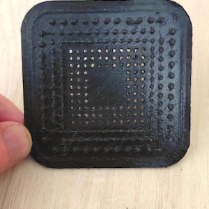 May include: A square black object with rounded corners featuring a textured surface with a grid of small raised dots arranged in concentric square patterns. The object has a flat, matte finish and a distinct layered appearance from 3D printing.