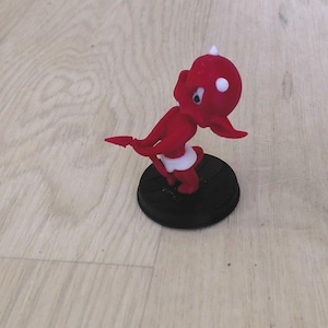 Peut inclure: Une petite figurine rouge représentant un personnage de diable de dessin animé avec une couche blanche, des cornes blanches et une tache blanche sur la tête. Le personnage a une queue pointue et est fixé sur une base noire circulaire.