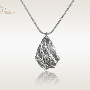 Puede incluir: Un collar de plata con un colgante texturizado de forma irregular. El colgante tiene un diseño en relieve que se asemeja a una letra hebrea.