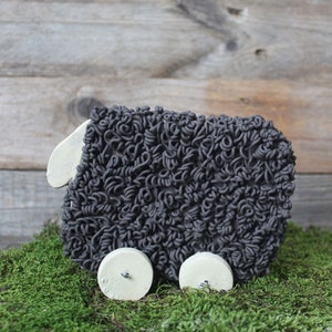 May include: A black sheep toy with a curly wool texture and white wheels. The toy is sitting on a bed of green moss.