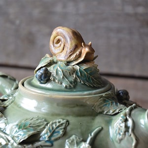 Könnte beinhalten: Eine grüne Teekanne aus Keramik mit einer Schnecke auf dem Deckel. Die Teekanne ist mit Blättern und Beeren verziert.