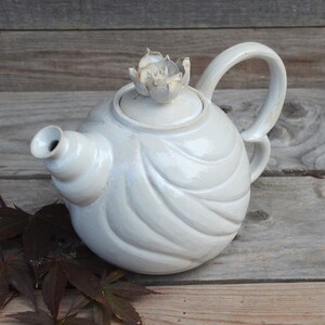 May include: White ceramic teapot with a flower-shaped lid and a wavy design. The teapot is sitting on a wooden surface.