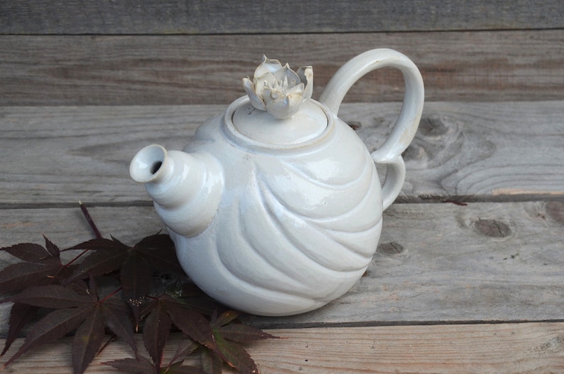 May include: White ceramic teapot with a flower-shaped lid and a wavy design. The teapot is sitting on a wooden surface.