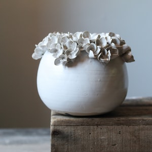 May include: White ceramic planter with a raised, textured rim of white flowers. The planter is sitting on a wooden surface.