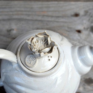 May include: White ceramic teapot with a flower-shaped lid and a decorative design on the side.