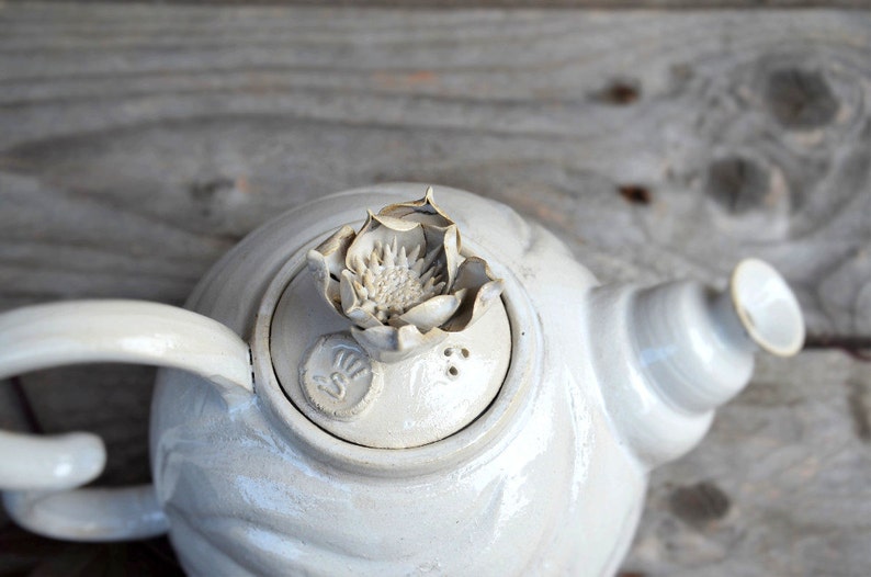 May include: White ceramic teapot with a flower-shaped lid and a decorative design on the side.