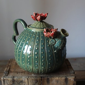 May include: A ceramic teapot in the shape of a cactus, with a green glaze and a textured finish. The teapot has a handle, a spout, and a lid adorned with a red flower. It is displayed on a wooden surface.