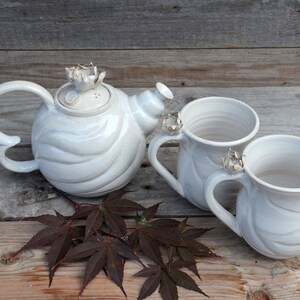 May include: White ceramic teapot and two mugs with floral accents. The teapot has a swirling design and the mugs have a smooth finish. The set is displayed on a wooden surface with brown leaves.