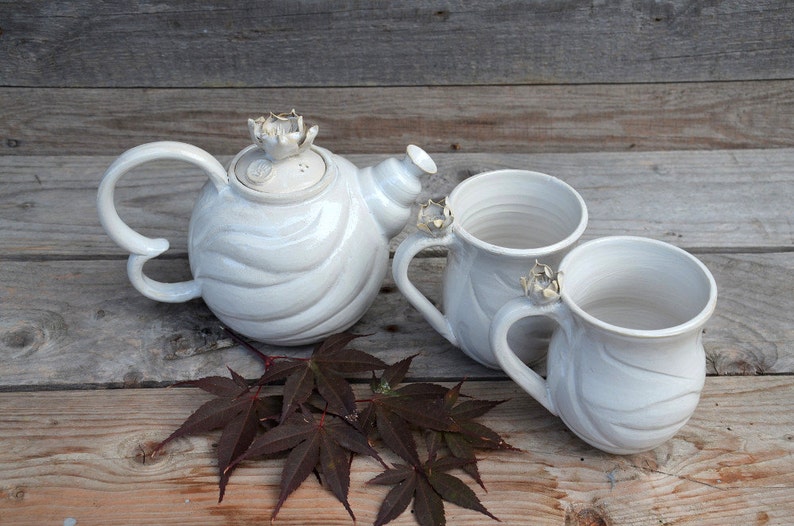 May include: White ceramic teapot and two mugs with floral accents. The teapot has a swirling design and the mugs have a smooth finish. The set is displayed on a wooden surface with brown leaves.