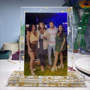 May include: A clear resin photo frame with gold leaf accents. The frame holds a color photograph of four people. The frame has a rectangular shape with a base for standing. The gold leaf is embedded throughout the resin.