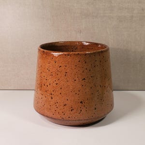 May include: A handmade, speckled ceramic mug in a warm, earthy brown hue. The mug has a slightly tapered shape, with a wider top and a narrower base. The surface is covered in small, dark brown speckles, giving it a rustic appearance. The interior is a darker shade of brown.