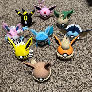 May include: A collection of nine colorful character figures, each with unique designs and features. The figures are arranged on a carpeted surface, showcasing a variety of colors including pink, blue, yellow, and black. The figures appear to be from a popular animated series.