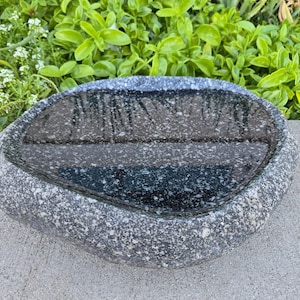 May include: A grey, oval-shaped stone bird bath. The interior is dark and appears to hold water. The bird bath sits on a concrete surface, with green foliage and white flowers in the background.