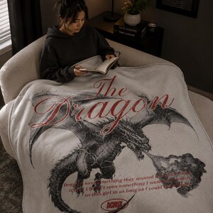 May include: A light gray blanket with a large dragon illustration in black and red. The text "The Dragon" is above the dragon, and other text is below. The blanket is draped over a person.