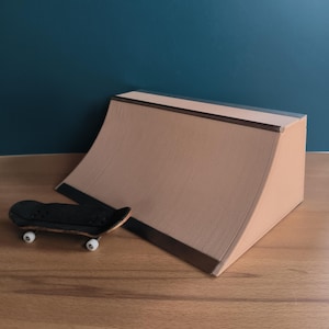 Könnte beinhalten: Ein kleines schwarzes Skateboard mit weißen Rädern neben einer beigefarbenen Rampe. Die Rampe hat eine gebogene Oberfläche und eine flache Oberseite mit dunkler Verkleidung. Der Hintergrund ist eine dunkelblaue Wand und eine Holzoberfläche. Die Rampe ist etwa 20 cm hoch.