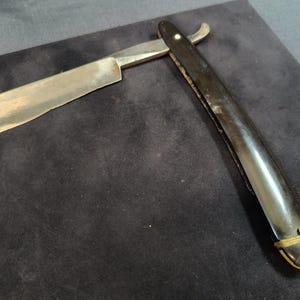 May include: A vintage straight razor with a black handle and a silver blade. The handle has a gold band and a decorative design.