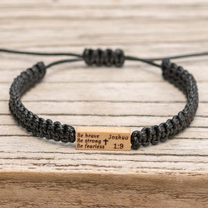 May include: A black cord bracelet featuring a rectangular wooden bead engraved with the text Be brave Be strong Be fearless Joshua 1:9 and a small cross symbol. The bracelet has an adjustable sliding knot closure made of black cord.