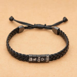 Op de afbeelding: Zwart gevlochten leren armband met verstelbare koordsluiting. De armband heeft vier zwarte kralen met de namen "Alex" en "Sara", een babyvoetafdruk en een hartsymbool. Een modieus accessoire.