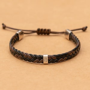 Könnte beinhalten: Ein geflochtenes Armband aus abwechselnd dunkelbraunen und schwarzen Ledersträngen. Das Armband verfügt über eine mittlere, silberfarbene Metallperle mit eingraviertem Dreieckssymbol, zwei silberfarbene Endkappen und einen verstellbaren Schiebeknoten.