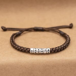 Op de afbeelding: Gevlochten bruine armband met zilveren kralen. De armband heeft een geweven ontwerp met verstelbare bruine koorden. De zilveren kralen hebben kleine zwarte stippen. Een stijlvol accessoire.