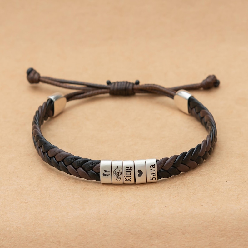 May include: A braided dark brown leather bracelet featuring five cylindrical silver-toned metal beads. The beads are engraved with a stick figure icon, a pinky promise hand gesture, the word King, a heart symbol, and the name Sara.