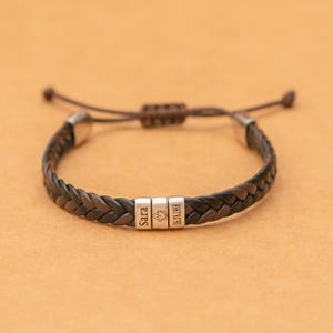 Könnte beinhalten: Ein geflochtenes Armband aus dunkelbraunen und schwarzen Ledersträngen. Das Armband verfügt über drei silberfarbene Metallperlen, die mit dem Namen Sara, einem kleinen Geistersymbol und dem Datum 06.08.1968 graviert sind. Es hat einen Schiebeknoten.