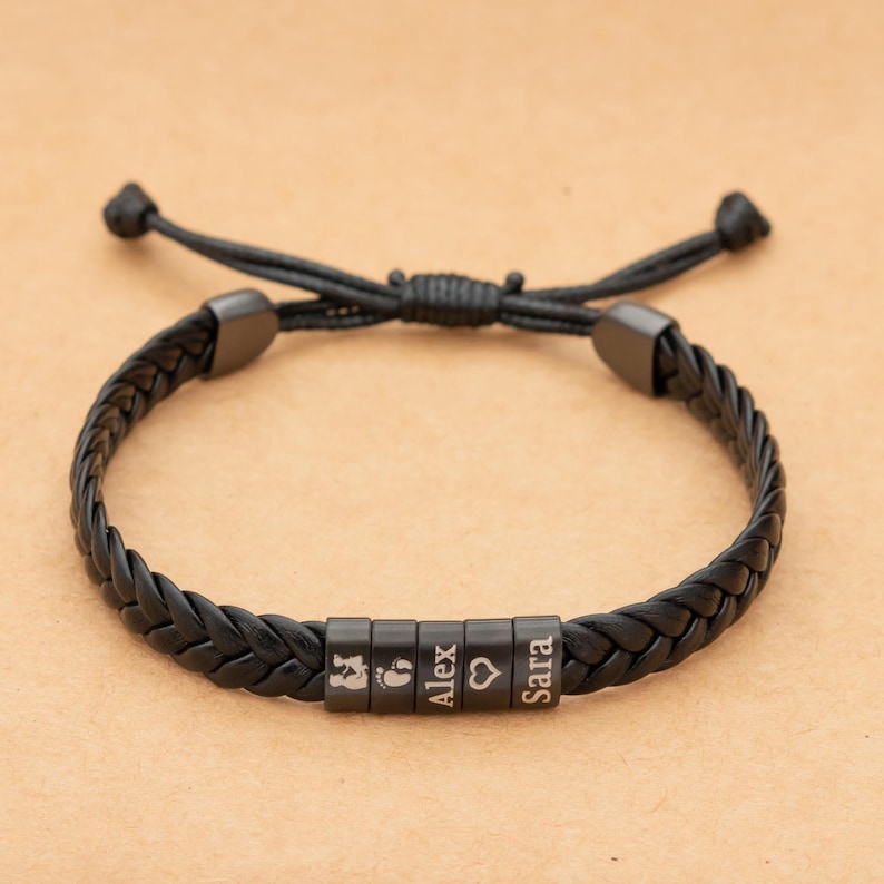 May include: A black braided cord bracelet featuring five cylindrical beads. The beads are engraved with a baby silhouette, a footprint, the name Alex, a heart symbol, and the name Sara. The bracelet has an adjustable sliding knot closure.