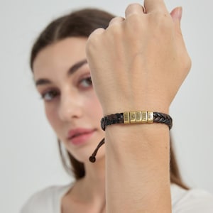 May include: A braided dark brown leather bracelet featuring five rectangular gold-toned metal beads. The beads are engraved with the text "Sara", a heart symbol, "Allen", "1989", and a small icon of two hands holding pinky fingers.