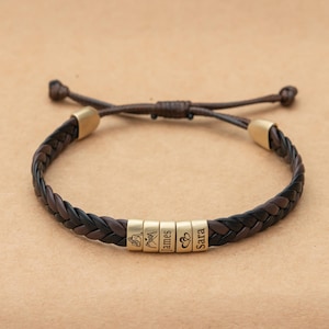 May include: A braided dark brown leather bracelet featuring five cylindrical gold-toned metal beads. The beads are engraved with a line drawing of two people, a mountain range, the name James, two hearts, and the name Sara, respectively.