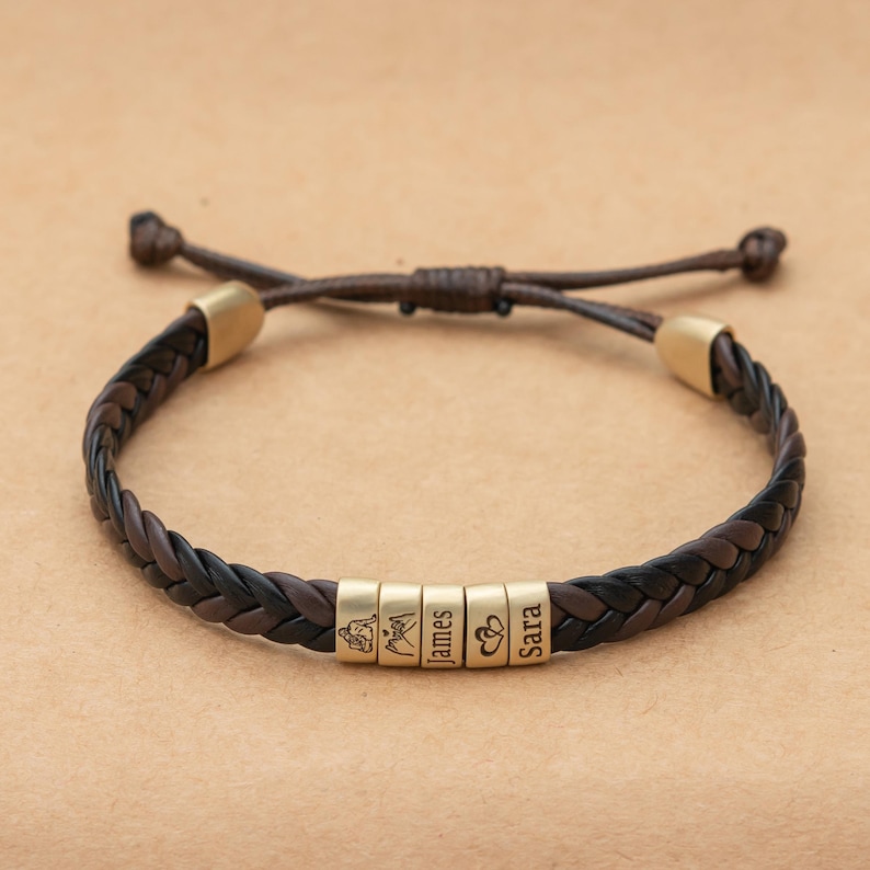 May include: A braided dark brown leather bracelet featuring five cylindrical gold-toned metal beads. The beads are engraved with a line drawing of two people, a mountain range, the name James, two hearts, and the name Sara, respectively.